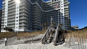 A multi-story hotel with balconies, a sandy beach in front, and a wooden fence.