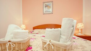 Une chambre avec deux fauteuils en osier et des serviettes, un lit avec une literie à motifs floraux, une tête de lit en bois, une table de chevet avec une lampe et un tableau encadré au mur.