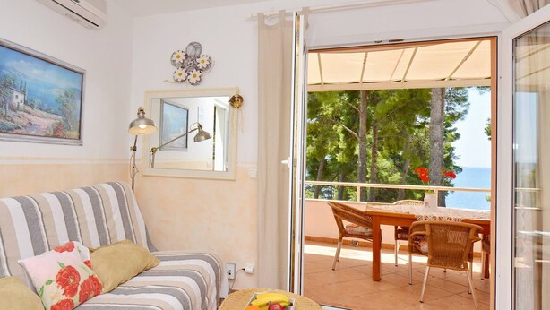 Une pièce chaleureuse avec un canapé à rayures, une petite table ronde sur laquelle sont disposés des fruits, un balcon avec une table et des chaises, et une vue sur la mer.