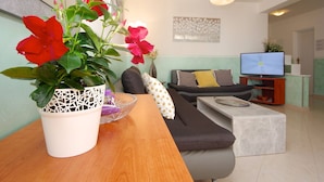 Una sala de estar con un sofá, una mesa de centro y un televisor. Hay una planta en maceta con flores rojas sobre una mesa de madera.