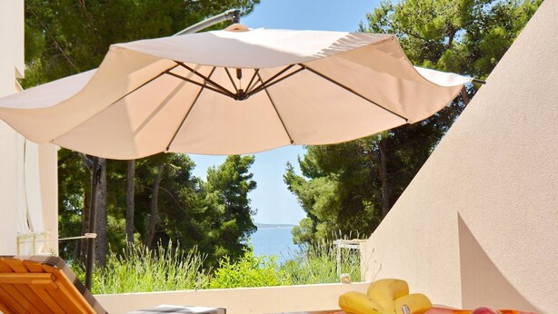 Una mesa de madera con un cuenco de fruta y un paraguas color beige en un balcón con vista a un cuerpo de agua y árboles.
