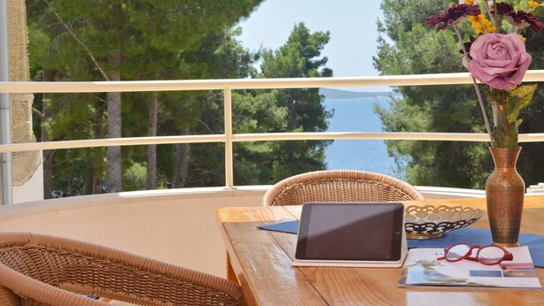 A balcony with a wicker chair, a wooden table with a tablet, a vase with flowers, and a view of the sea and trees. - Seafront Apartments Deveron - Duplex Two Bedroom Apartment with Balcony and Sea View (Jelsa)