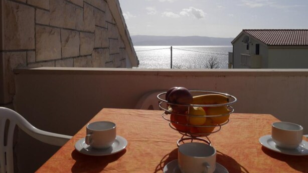 A balcony with a table set for two, a metal fruit basket, and a view of the sea and distant mountains.