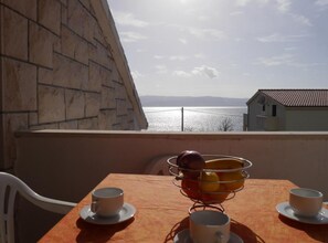 En balkon med et bord dækket til to, en fletkurv med frugt og udsigt over havet og fjerne bjerge.