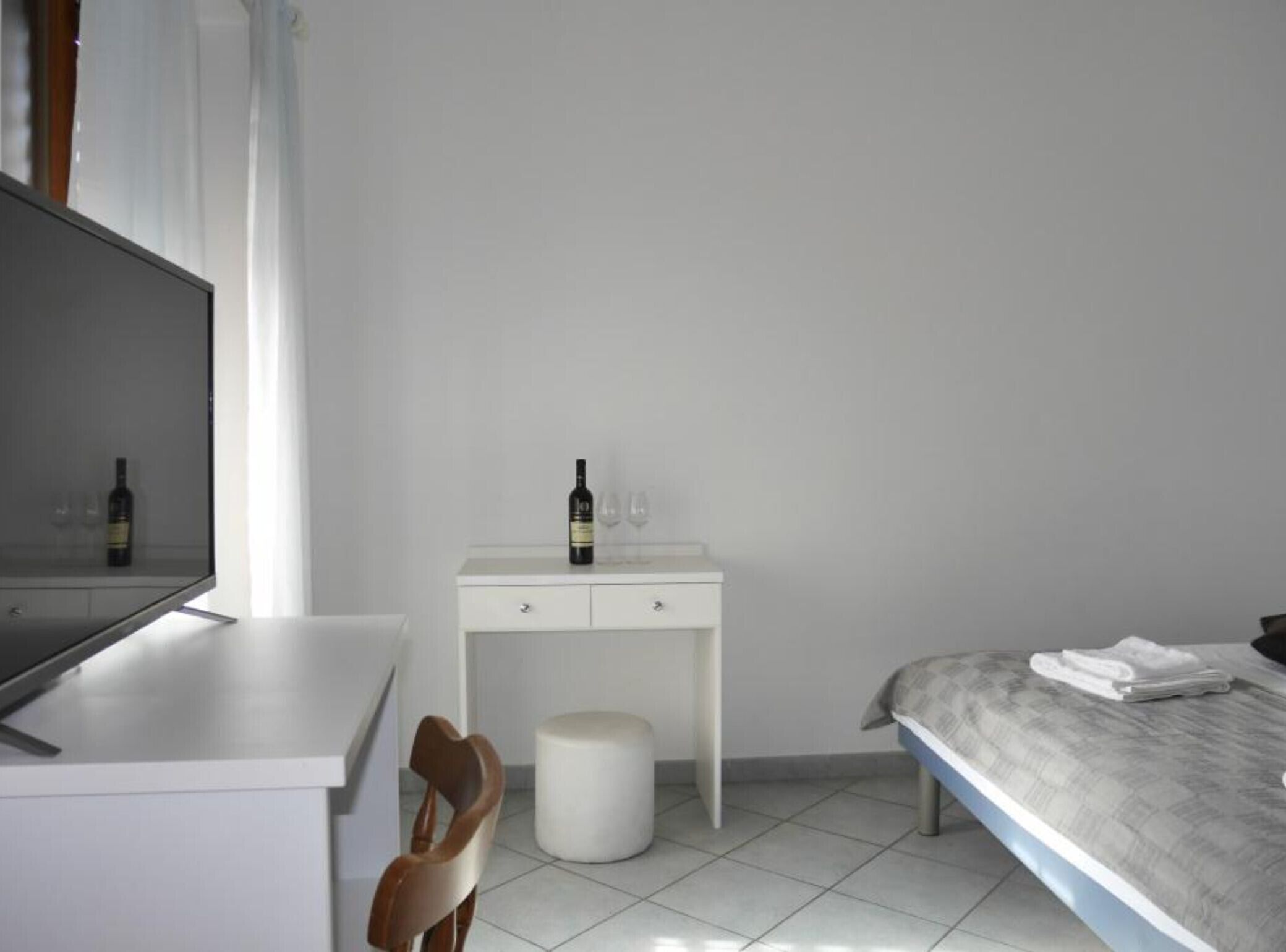 Habitación de hotel con cama, un pequeño escritorio blanco con una botella de vino y copas, una silla y un taburete redondo.