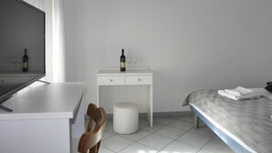 A hotel room with a bed, a small white desk with a bottle of wine and glasses, a chair, and a round stool.