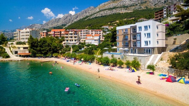 Un hôtel situé sur la côte, avec plusieurs étages, une plage au premier plan et des montagnes en arrière-plan.
