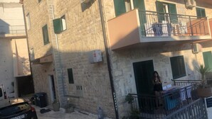 A two-story stone building with green shutters, a balcony with a metal railing, and a parked car in the foreground.
