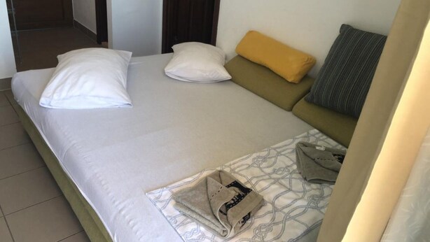 A single bed with white linens, a yellow pillow, and a gray pillow. There is a folded towel and a patterned blanket on the bed.
