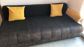 A dark-colored sofa with two yellow cushions against a plain wall, placed on a tiled floor.