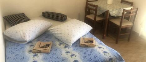 A small room with a bed, two pillows, a striped pillow, a small table with a tablecloth, and two chairs.
