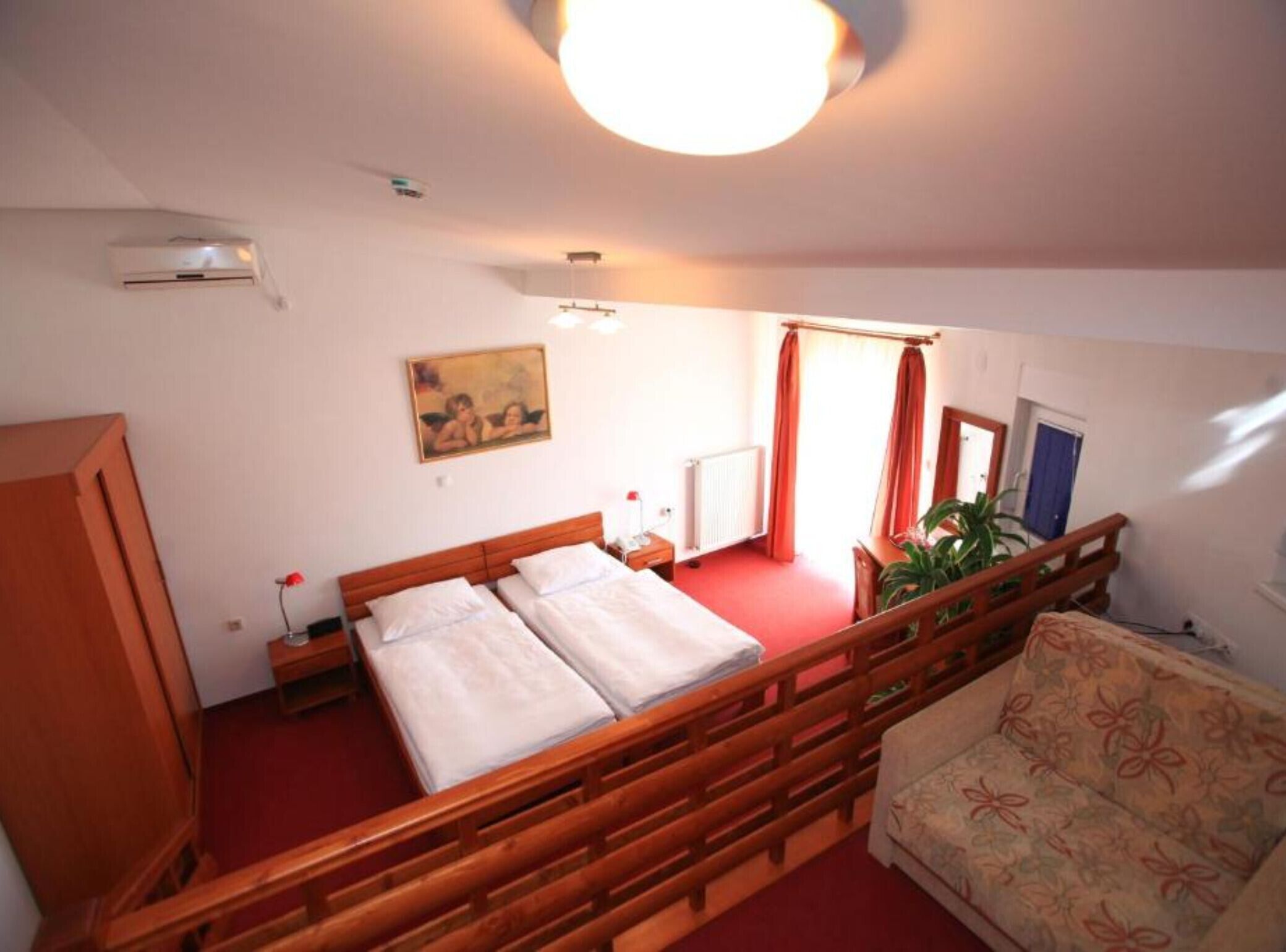 A bedroom with a wooden bed, a nightstand, a desk, a chair, a sofa, a radiator, a painting, and a window with curtains.