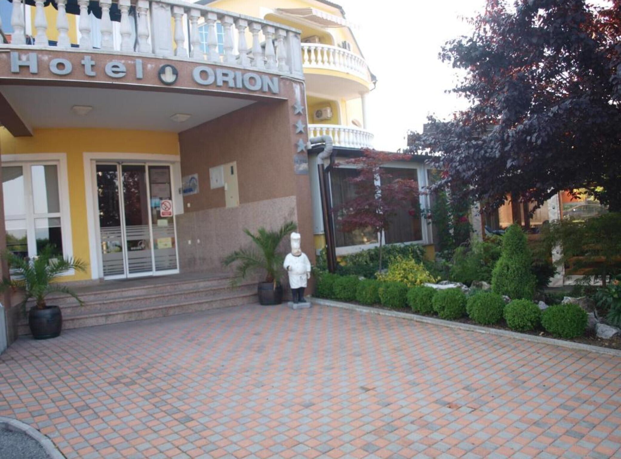 A hotel entrance with a sign reading "Hotel Orion," a statue of a chef, and a paved walkway.