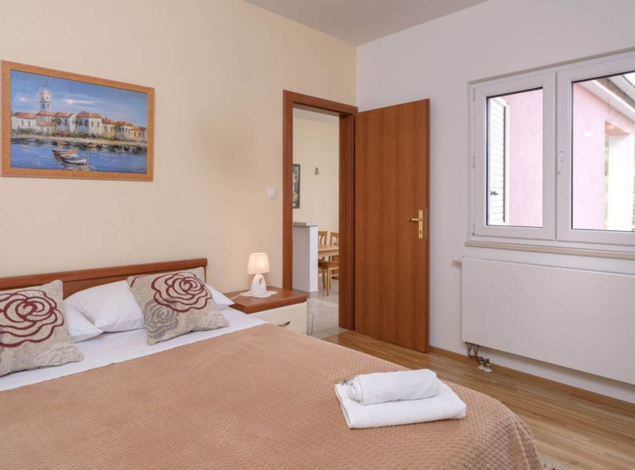 Quarto de hotel com cama, mesa de cabeceira, luminária, pintura de uma paisagem costeira, porta de madeira e janela com vista para um edifício.