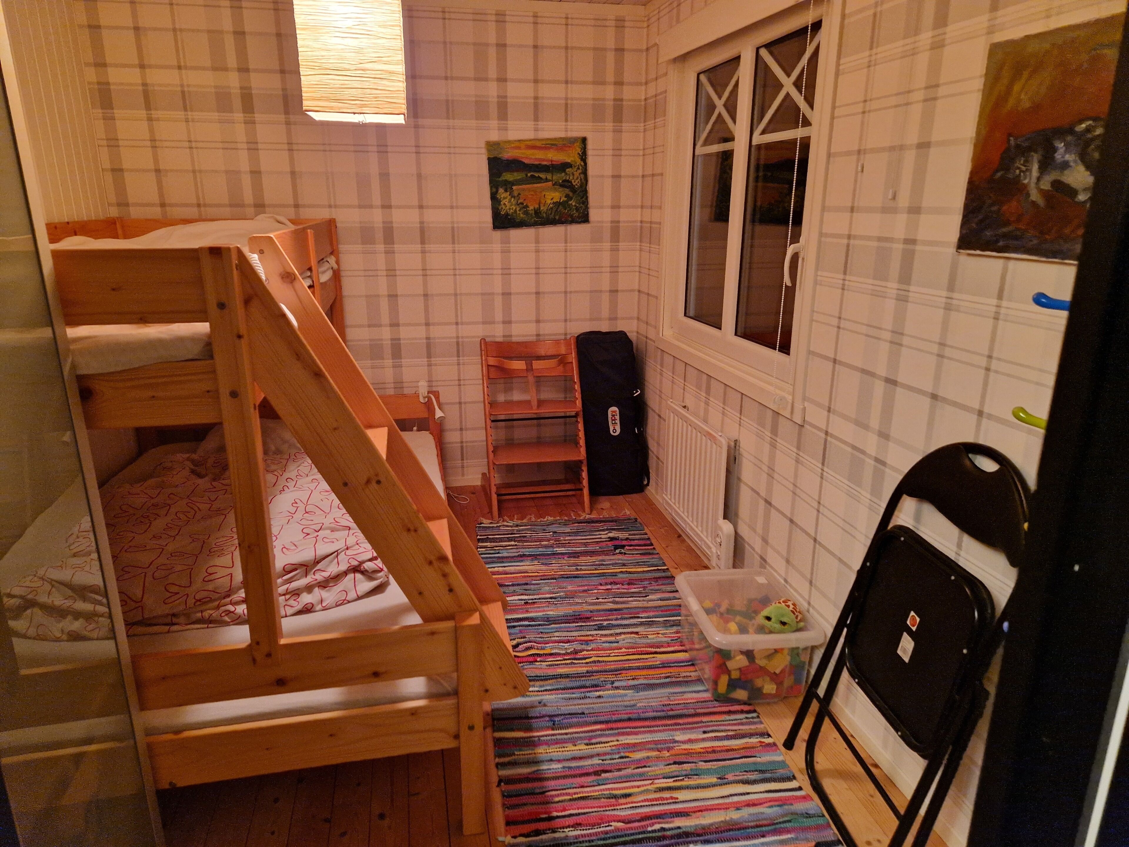 Een kamer met een stapelbed, een raam, een radiator, een stoel en een kleurrijk vloerkleed.