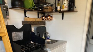 A compact kitchen with a gas stove, a sink, and a trash bin. There is a wooden ladder leaning against the wall, and a small shelf above the stove holding kitchen utensils. - The Greenhouse - New River Gorge National Park (Rainelle)