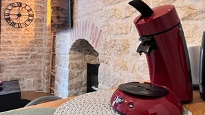 A red coffee maker on a woven placemat in a stone-walled room with a clock and a fireplace.