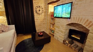 A cozy living room with a stone wall, a fireplace, a flat-screen TV, a clock, and a black ottoman with decorative items.