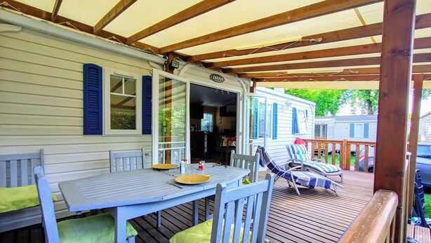 A covered outdoor deck with a dining table and chairs, a lounge area with a striped cushion, and a view of a mobile home in the background. - FULLY EQUIPPED MOBIL HOME with large 30 M2 terrace and small garden. (Saint-Augustin)