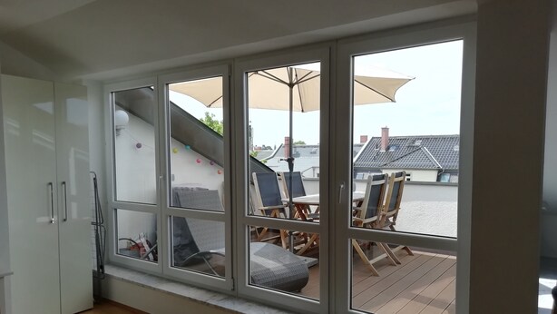 A room with a large window overlooking a deck area with outdoor furniture and umbrellas. - Duplex apartment over 2 levels with roof terrace near the Elbe in Dresden-Blasewitz (Dresden)
