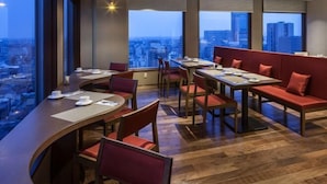 Ein modernes Restaurant mit hohen Tischen und rot gepolsterten Sitzplätzen, große Fenster mit Blick auf die Stadt.