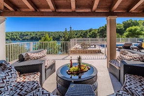A covered outdoor seating area with wicker furniture, a table with fruit, and a view of a lake and forest. - Luxury 4BR Seafront Villa with Heated Pool (Milna)