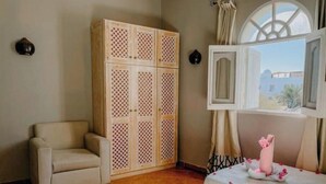 A room with a beige sofa, a wooden cabinet with a lattice design, a bed with a white and pink floral bedspread, and a window with a view of buildings. - Eel Garden Villas - mountain view beach side apt. (Dahab)
