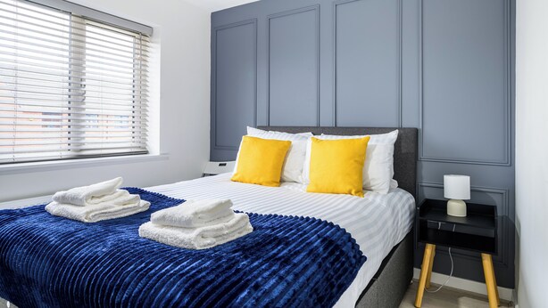 Ein modernes Schlafzimmer mit einem Bett, zwei gelben Kissen, einer blauen Decke und einem Nachttisch mit einer Lampe.