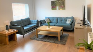 A modern living room with a grey sofa, a wooden coffee table, a small side table, a TV, and a refrigerator.