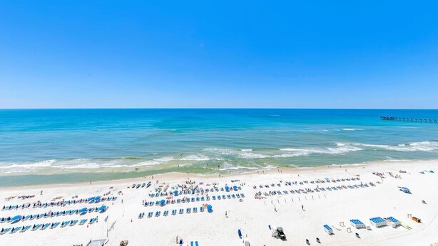 Ein Strand mit Reihen von Liegestühlen und Sonnenschirmen, einer in das Wasser führenden Mole und einem klaren, blauen Himmel.