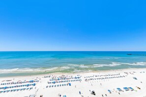A beach with rows of sun loungers and umbrellas, a pier extending into the water, and a clear blue sky. - Aqua Resort 1207 (Panama City Beach)