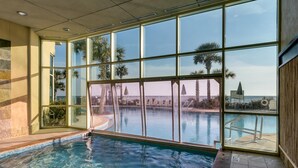 Une piscine intérieure avec des parois en verre transparent, un sol carrelé et une vue sur des palmiers et un bâtiment situé au loin.