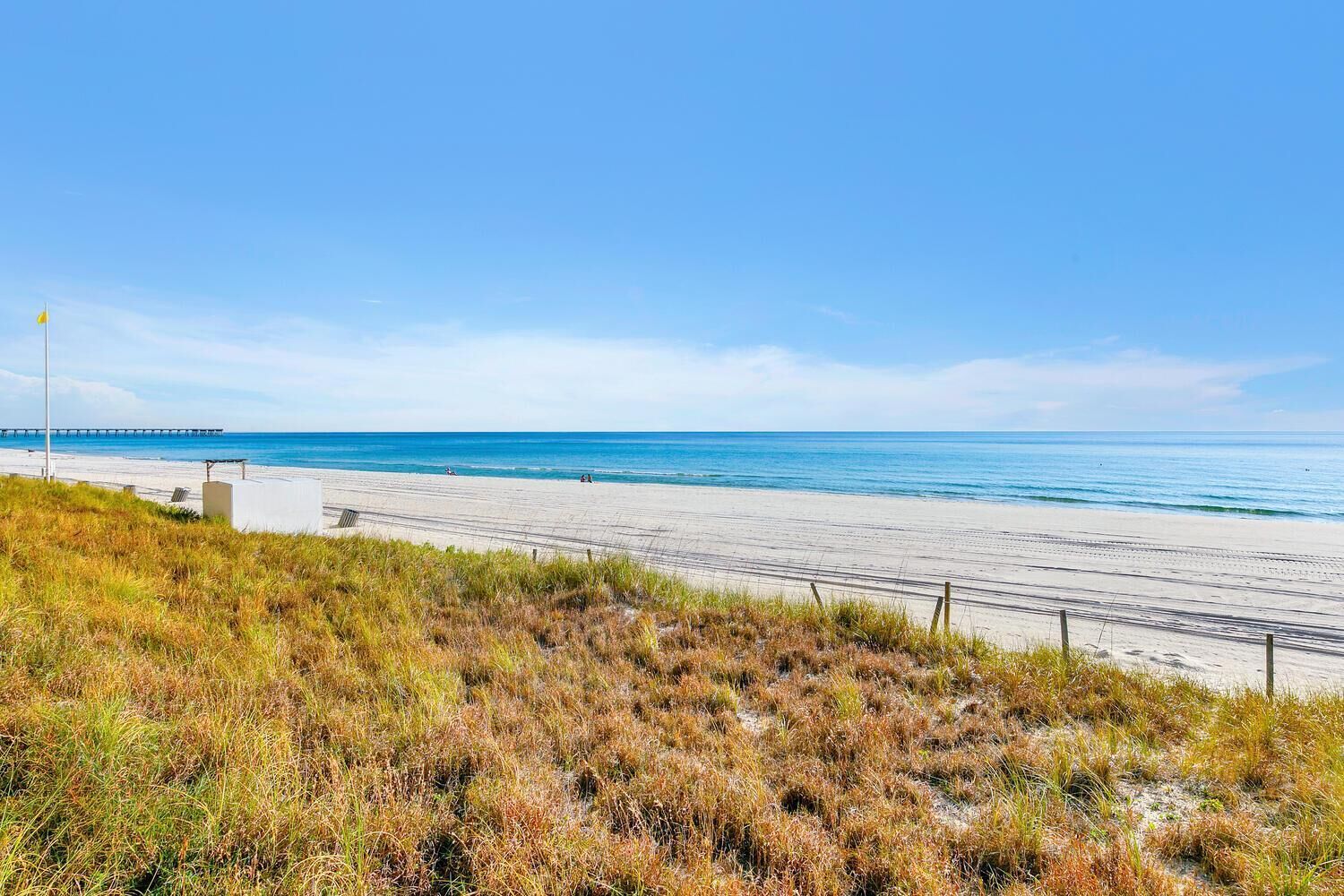 Uma praia com um píer ao longe, areia dourada e dunas gramadas.