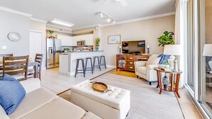 A modern living room with a sofa, ottoman, and a small table. There is a kitchen area with a refrigerator, microwave, and bar stools. A dining table with chairs is also visible. - Tidewater 913 (Panama City Beach)