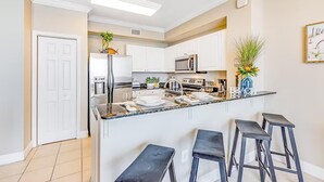 A modern kitchen with white cabinets, a central island with bar stools, a microwave, and a refrigerator. - Tidewater 913 (Panama City Beach)