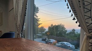 Una mesa de madera y una ventana con cortinas, con vistas a una calle con coches y árboles al atardecer.