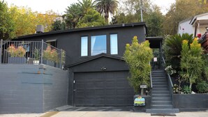Una casa moderna de dos pisos con un garaje negro, un pequeño jardín delantero con un árbol y un buzón, y una señal de calle que indica la dirección.