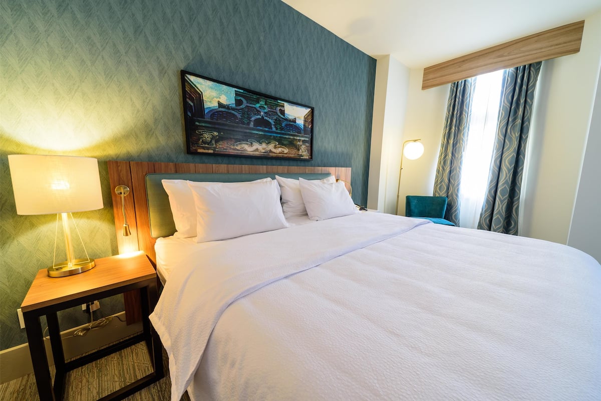 A hotel room with a large bed, a nightstand with a lamp, a painting on the wall, and a window with curtains.