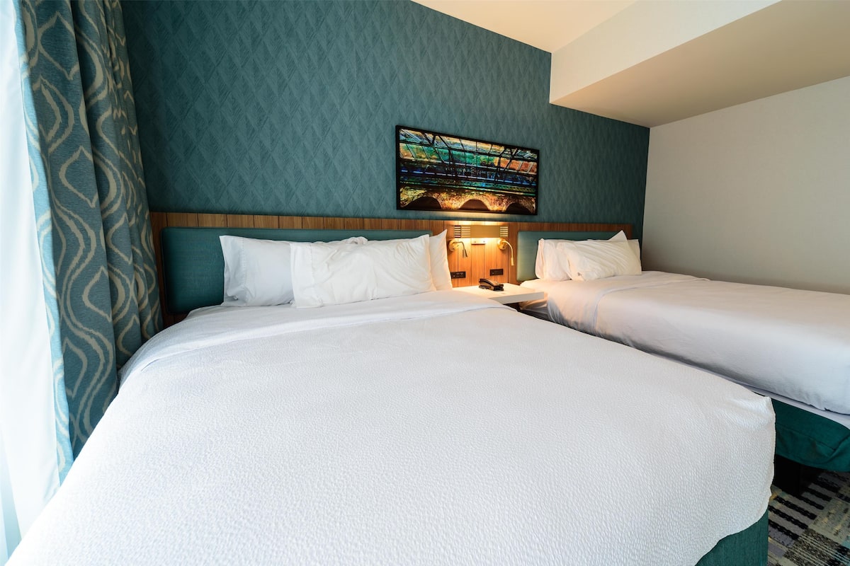 A hotel room with two beds, a nightstand, a wall-mounted artwork, and a patterned curtain.