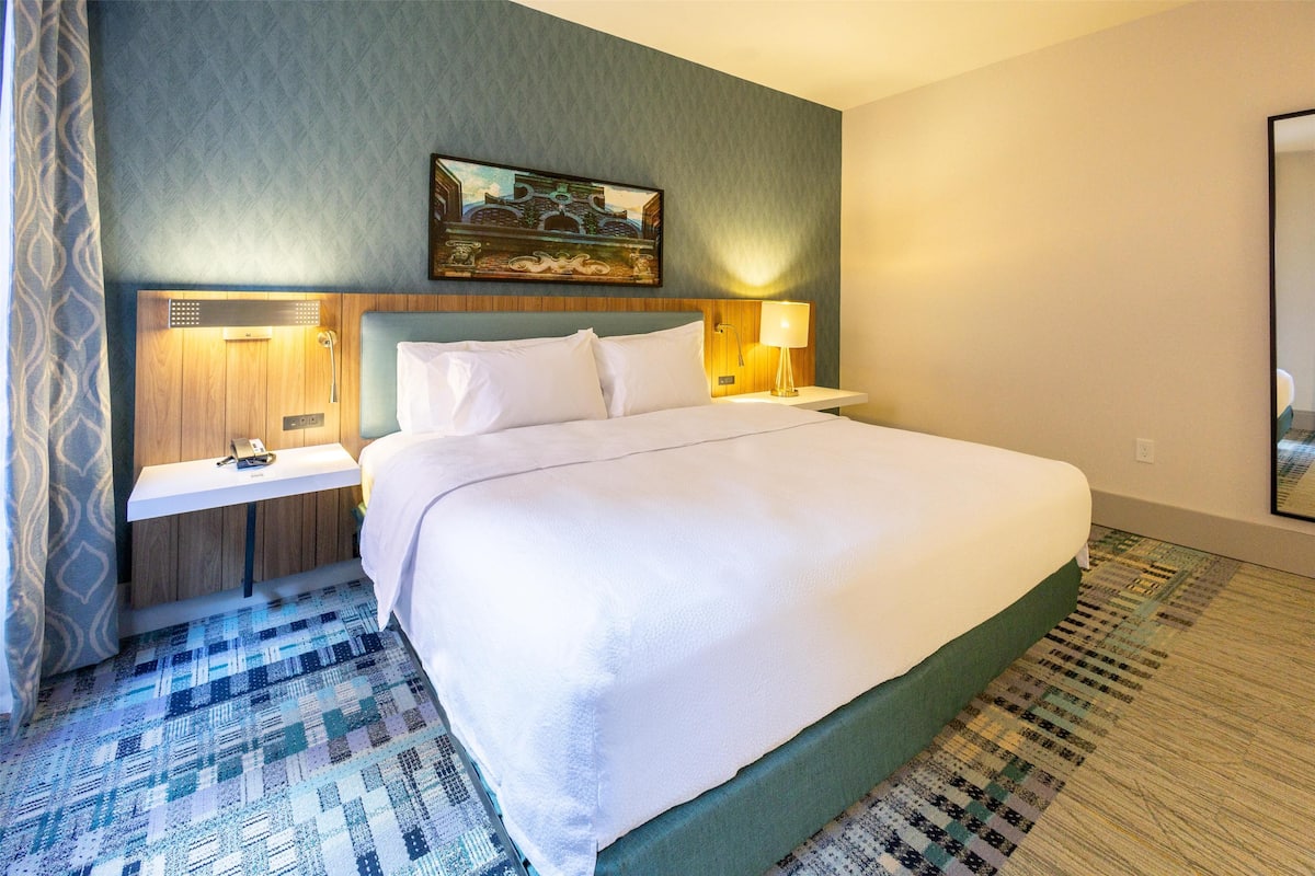 A hotel room with a large bed, a nightstand with a lamp, a painting on the wall, and a patterned carpet.