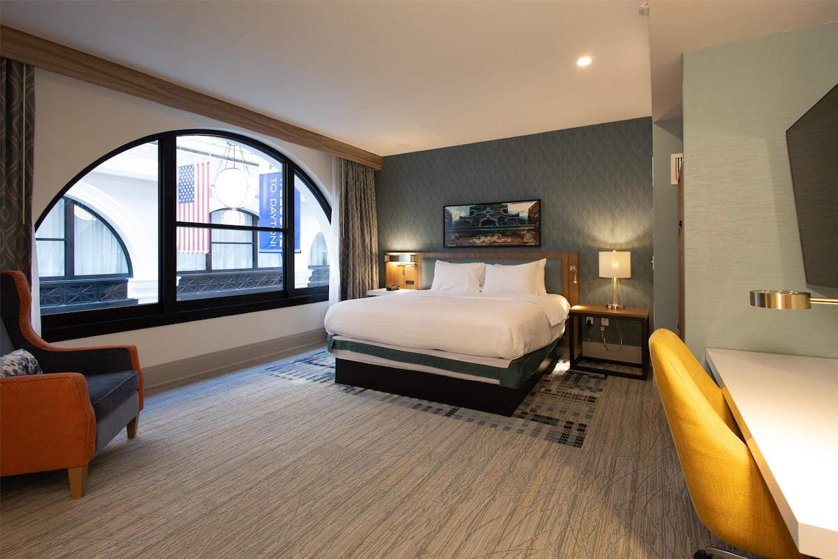 A hotel room with a large bed, a desk, a chair, and a window with a view of a building and an American flag.