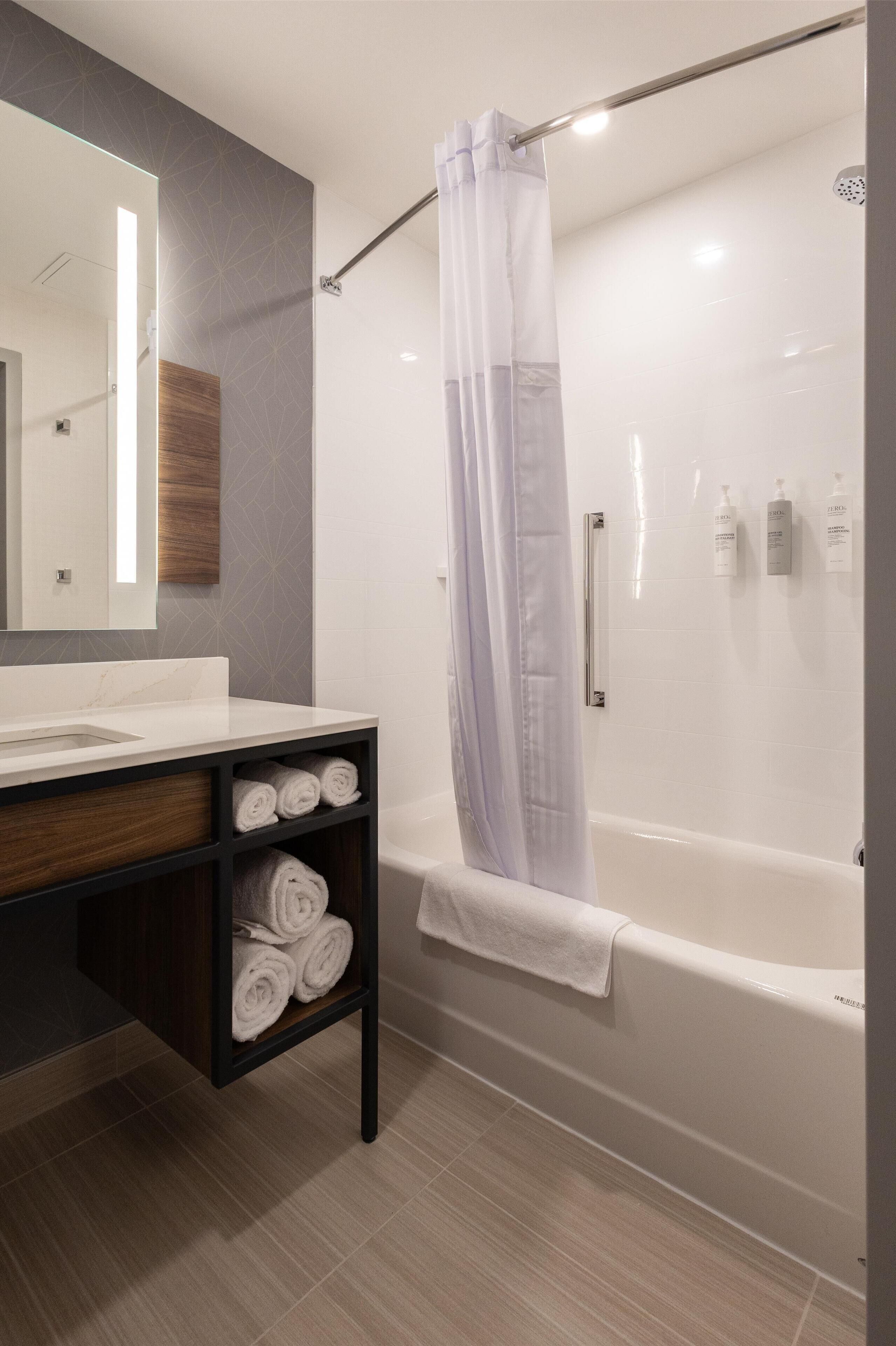 A modern bathroom with a white bathtub, a glass shower enclosure, a wooden vanity with a mirror, and neatly rolled towels.