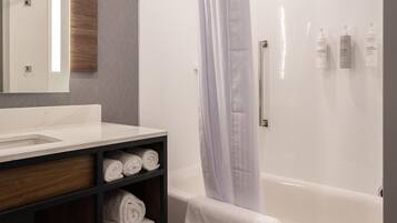 A modern bathroom with a white bathtub, a glass shower enclosure, a wooden vanity with a mirror, and neatly rolled towels.