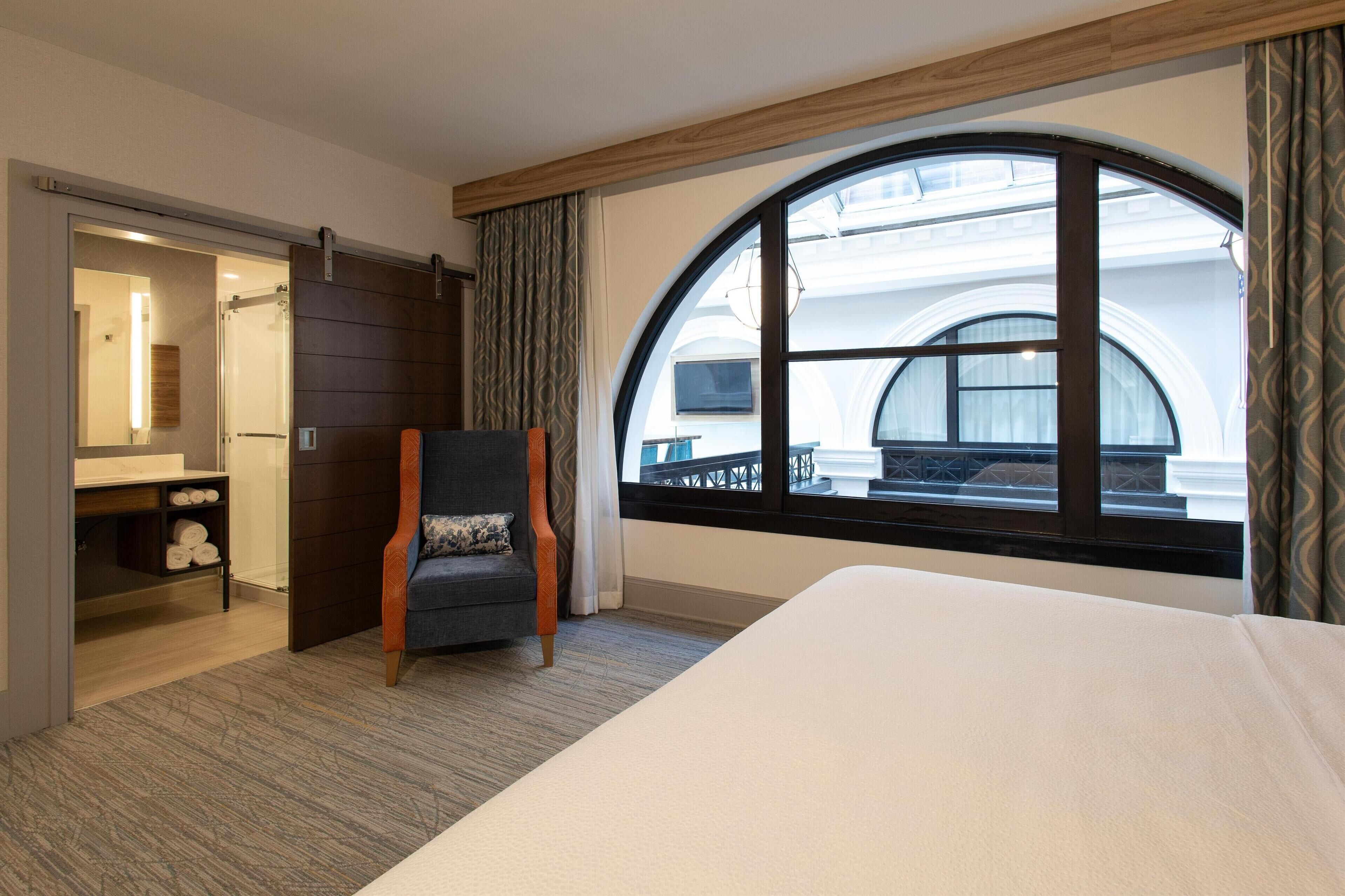 Hotellihuone, jossa on suuri kaarikkuna, sänky, tuoli, peilillä varustettu meikkipöytä ja erillinen vaatehuone.
