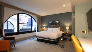Habitación de hotel con una cama grande, un escritorio, una silla y un ventanal con vistas a un edificio y una bandera estadounidense.
