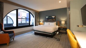 Habitación de hotel con una cama grande, un escritorio, una silla y un ventanal con vistas a un edificio y una bandera estadounidense.