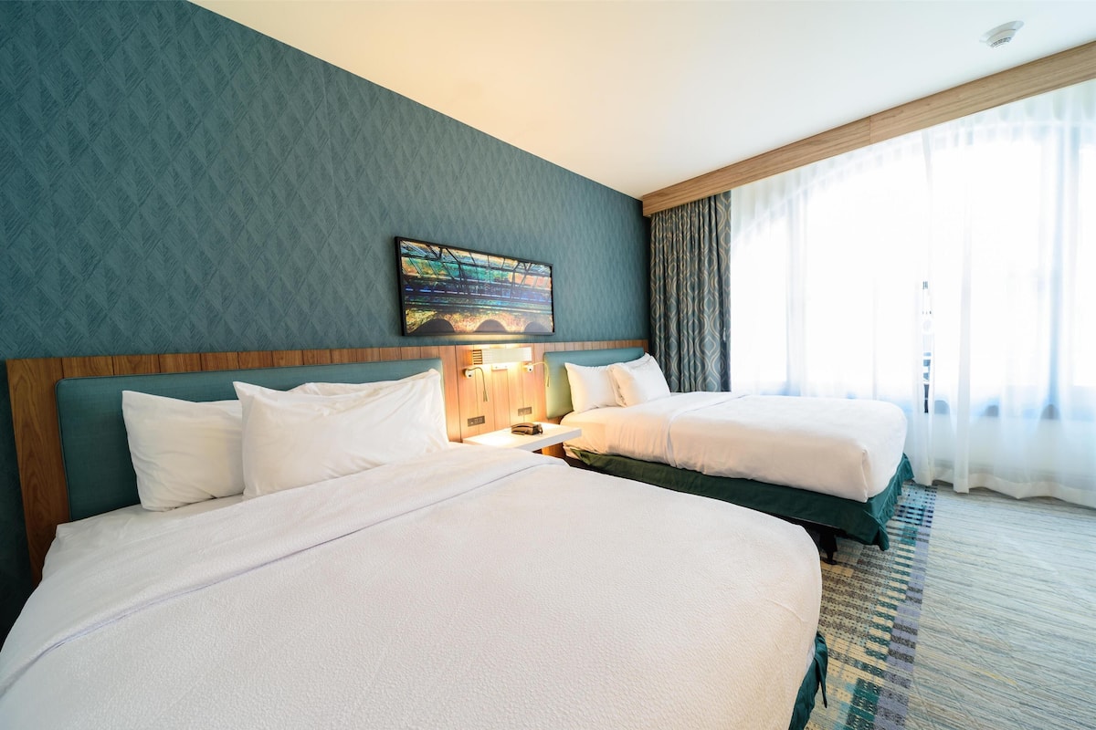 A hotel room with two beds, a large window with curtains, a painting on the wall, and a wooden headboard.