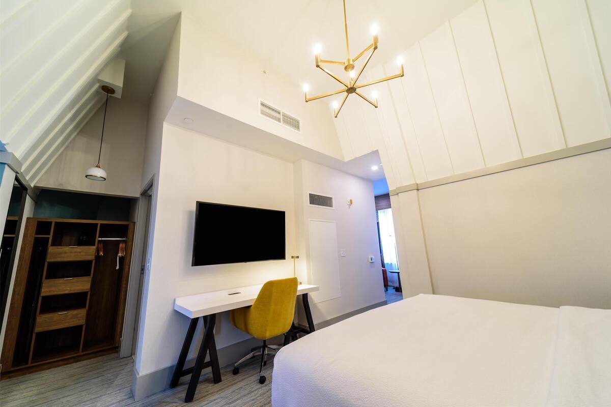 A modern hotel room with a bed, a desk with a chair, a TV on the wall, and a wardrobe.