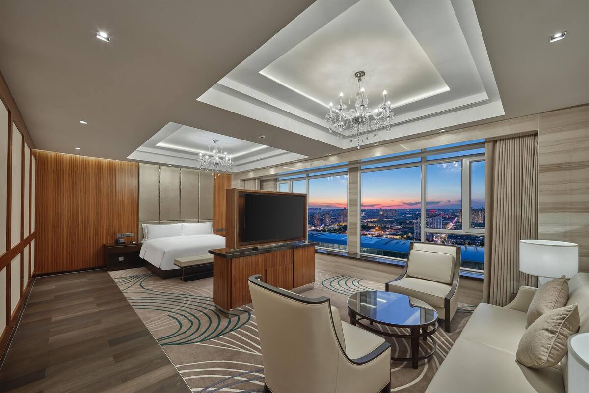 Ein modernes Hotelzimmer mit einem großen Bett, einem Fernseher, einer Sitzecke mit Glastisch und Blick auf die Stadt durch große Fenster.