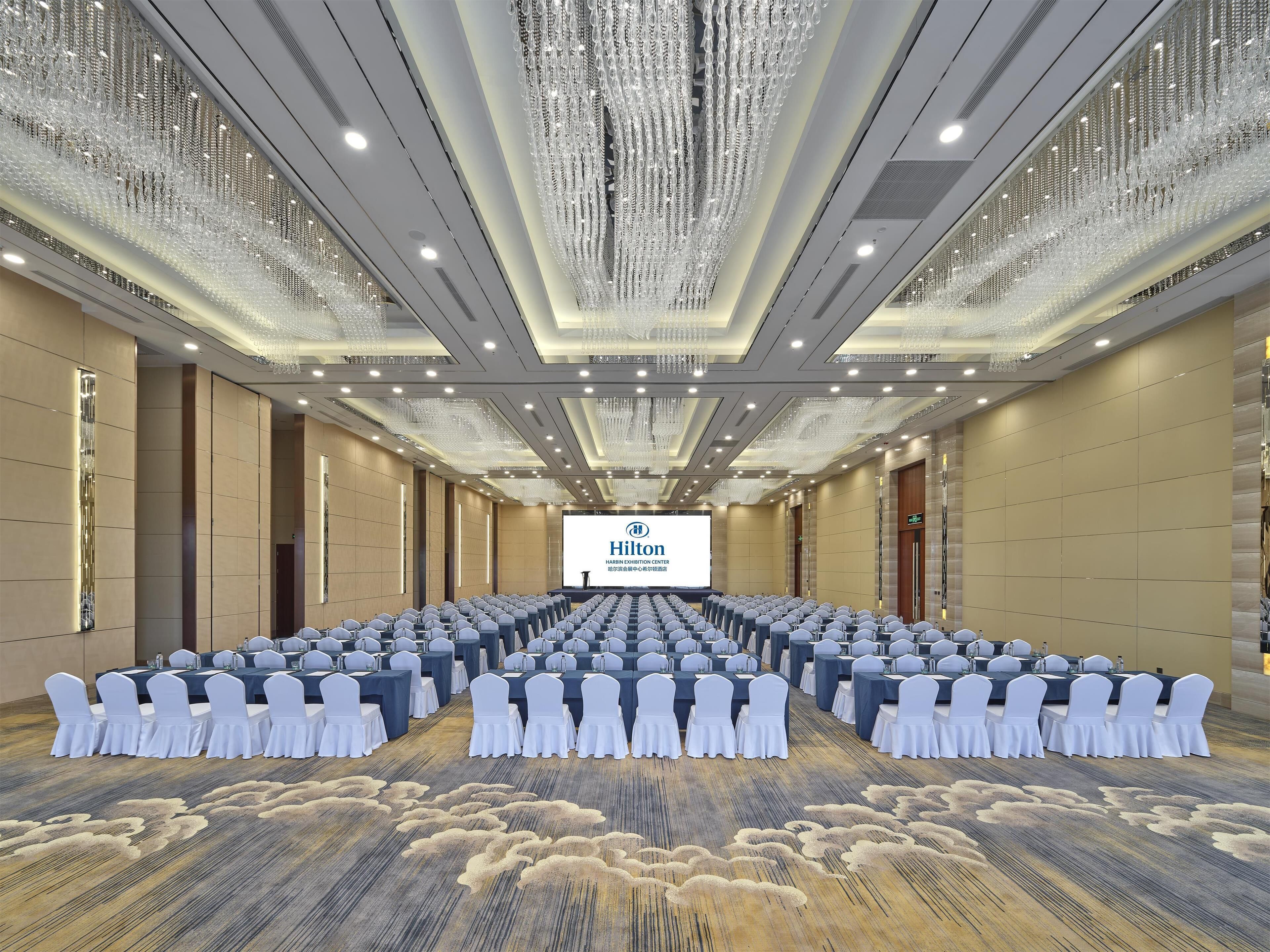 A large conference room with rows of tables and chairs, a large screen displaying 'Hilton', and a ceiling with hanging crystal chandeliers.